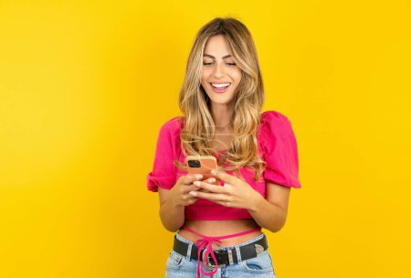 Foto de Mujer rubia joven que usa top rosa sobre fondo amarillo usando el teléfono móvil chateando en tiempo libre - Imagen libre de derechos