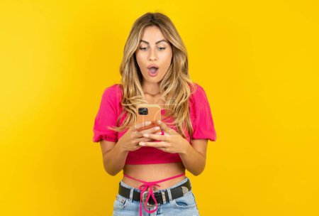 Foto de Mujer joven rubia sorprendida que usa top rosa sobre fondo amarillo usando un teléfono inteligente leyendo noticias de redes sociales, o un correo electrónico importante - Imagen libre de derechos