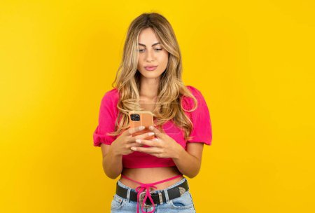 Photo for Focused blonde young woman wearing pink crop top on yellow background use smartphone reading social media news or important e-mail - Royalty Free Image