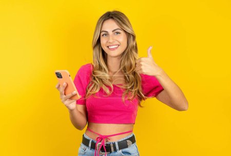 Photo for Portrait of blonde young woman wearing pink crop top on yellow background using and texting with smartphone with big smile doing ok sign, thumb up with finger, excellent sign - Royalty Free Image