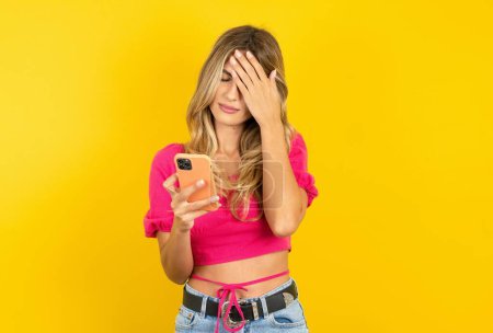 Photo for Beautiful blonde young woman wearing pink crop top on yellow background looking at smartphone feeling sad holding hand on face. - Royalty Free Image