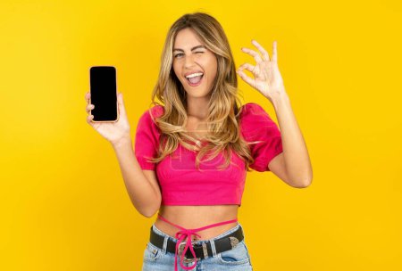 Photo for Beautiful blonde young woman wearing pink crop top on yellow background holding in hand smartphone showing ok sign - Royalty Free Image