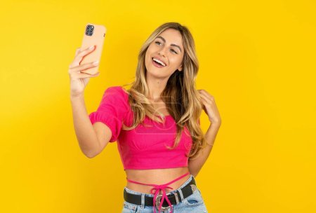 Photo for Beautiful blonde young woman wearing pink crop top on yellow background smiling and taking a selfie ready to post it on her social media. - Royalty Free Image