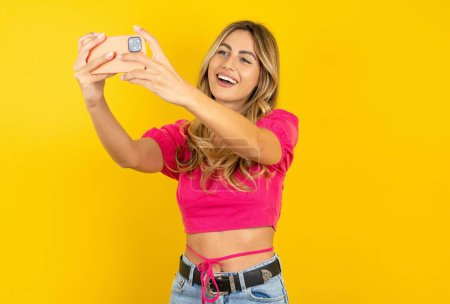 Photo for Beautiful blonde young woman wearing pink crop top on yellow background taking a selfie to post it on social media or having a video call with friends. - Royalty Free Image