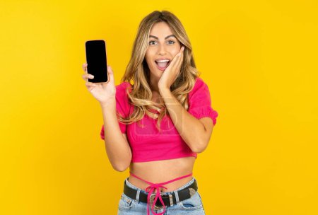 Photo for Impressed blonde young woman wearing pink crop top on yellow background with smartphone in hand touches face with palm - Royalty Free Image