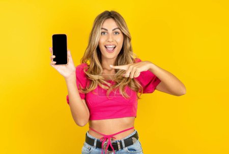 Téléchargez les photos : Joyeux blond jeune femme portant un haut de culture rose sur fond jaune tenant dans les mains cellule montrant écran noir - en image libre de droit