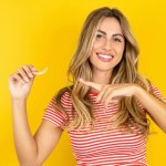 Beautiful blonde young woman wearing striped t-shirt over yellow studio background holding an invisible aligner and pointing at it. Dental healthcare and confidence concept. 