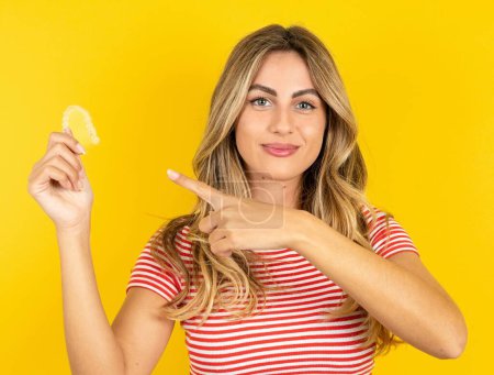 Téléchargez les photos : Jeune belle femme blonde portant un t-shirt rayé sur fond de studio jaune tenant un aligneur invisible et le pointant du doigt. Concept de soins dentaires et de confiance. - en image libre de droit
