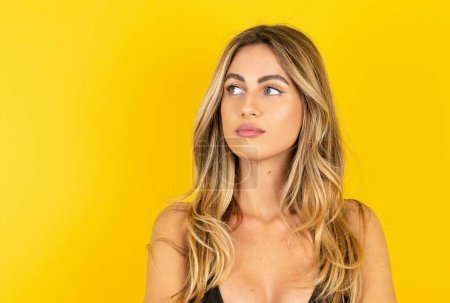 Photo for Close up portrait of charming woman wearing bikini over yellow background with pensive expression thinks what to do - Royalty Free Image