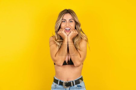 Photo for Happy young blonde woman wearing bikini over yellow background keeps fists on cheeks smiles broadly and has positive expression being in good mood - Royalty Free Image