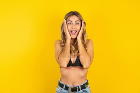 Foto de Hermosa joven rubia con palmas en la cara con bikini sobre fondo amarillo agradable aspecto alegre, feliz reacción - Imagen libre de derechos