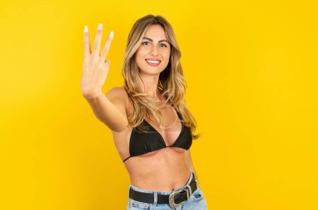 Foto de Hermosa mujer rubia joven con bikini sobre fondo amarillo sonriendo y mirando amigable, mostrando el número tres o tercero con la mano hacia adelante, cuenta atrás - Imagen libre de derechos