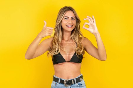 Téléchargez les photos : Positif jeune femme blonde portant un bikini sur fond jaune montrant pouces vers le haut et ok signes - en image libre de droit