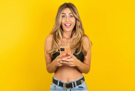 Foto de Hermosa mujer rubia joven con bikini sobre fondo amarillo sostiene el teléfono móvil en las manos y se alegra de las noticias positivas, utiliza celular moderno - Imagen libre de derechos