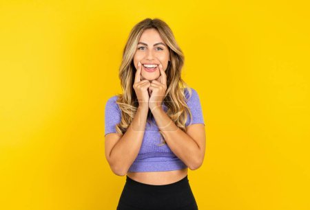 Téléchargez les photos : Heureuse femme blonde portant des vêtements de sport sur fond de studio jaune avec sourire aux dents, garde les index près de la bouche, les doigts pointés et forçant sourire joyeux - en image libre de droit