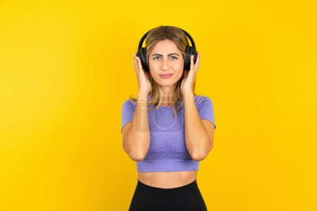 Téléchargez les photos : Sérieusement mécontente jeune femme dans les écouteurs portant des vêtements de sport sur fond jaune studio écoute de la musique et semble perplexe à la caméra - en image libre de droit