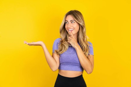 Foto de Mujer joven positiva que usa ropa deportiva sobre el fondo del estudio amarillo anuncia promoción y toca los dientes de los dedos - Imagen libre de derechos