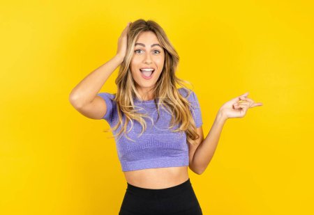 Photo for Surprised young woman wearing sportswear over yellow studio background pointing at empty space and holding hand on head - Royalty Free Image