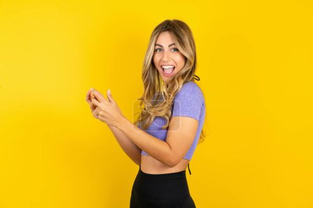 Photo for Cheerful young woman wearing sportswear over yellow studio background using gadget while playing network game - Royalty Free Image