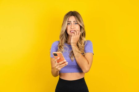 Téléchargez les photos : Peur jeune femme portant des vêtements de sport sur fond jaune studio tenant téléphone et ongles mordants - en image libre de droit