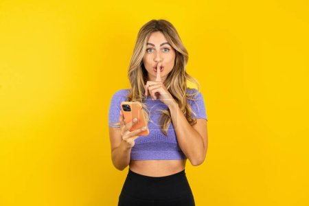 Photo for Young beautiful blonde woman wearing sportswear over yellow studio background holding modern gadget ask not tell secrets - Royalty Free Image