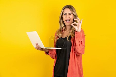 Photo for Attractive cheerful skilled young beautiful blonde woman standing over yellow studio background using laptop showing ok-sign winkin - Royalty Free Image