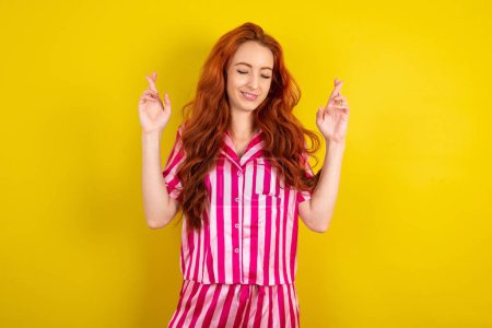 Téléchargez les photos : Femme aux cheveux roux portant un pyjama rose sur fond jaune studio a grand espoir, croise les doigts, croit en la bonne fortune, sourit largement. Concept de personnes et de souhaits - en image libre de droit