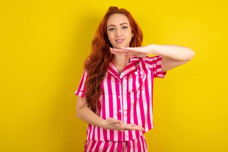 Photo for Red haired woman wearing pink pyjama over yellow studio background gesturing with hands showing big and large size sign, measure symbol. Smiling looking at the camera. - Royalty Free Image