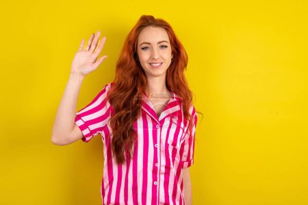 Téléchargez les photos : Femme aux cheveux roux portant un pyjama rose sur fond jaune studio saluant joyeusement et souriant, geste de bienvenue amical. - en image libre de droit