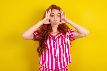Photo for Red haired woman wearing pink pyjama over yellow studio background with scared expression, keeps hands on head, jaw dropped, has terrific expression. Omg concept - Royalty Free Image