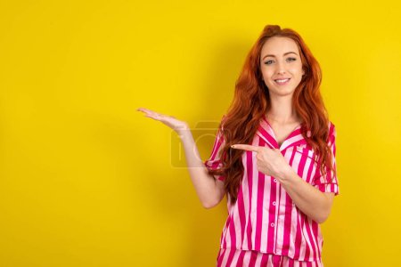 Téléchargez les photos : Positif femme aux cheveux rouges portant pyjama rose sur fond jaune studio promotion et index pointu sur copyspace maintenez la main démontrer offre annonces promo - en image libre de droit