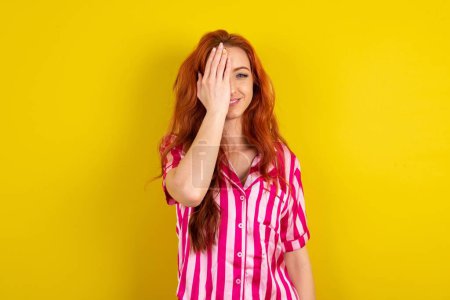 Téléchargez les photos : Jeune femme aux cheveux rouges portant un pyjama rose sur fond jaune studio couvrant un œil avec sa main, sourire confiant sur le visage et émotion surprise. - en image libre de droit