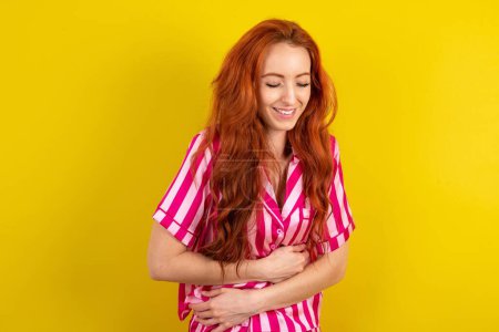 Foto de Joven mujer pelirroja con pijama rosa sobre fondo de estudio amarillo sonriendo y riendo en voz alta porque divertida broma loca con las manos en el cuerpo. - Imagen libre de derechos