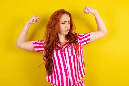 Foto de Joven mujer pelirroja con pijama rosa sobre fondo de estudio amarillo mostrando los músculos de los brazos sonriendo orgullosa. Concepto Fitness. - Imagen libre de derechos