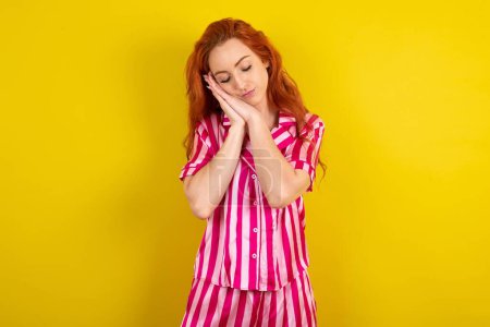 Foto de Mujer pelirroja vistiendo pijama rosa sobre fondo amarillo estudio durmiendo cansado soñando y posando con las manos juntas mientras sonríe con los ojos cerrados. - Imagen libre de derechos
