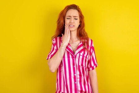 Foto de Mujer pelirroja con pijama rosa sobre fondo amarillo estudio tocando la boca con la mano con expresión dolorosa debido a dolor de muelas o enfermedad dental en los dientes. - Imagen libre de derechos
