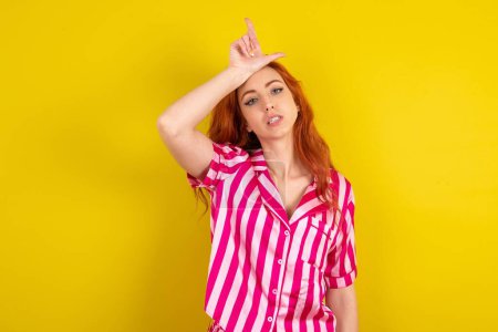 Photo for Red haired woman wearing pink pyjama over yellow studio background making fun of people with fingers on forehead doing loser gesture mocking and insulting. - Royalty Free Image