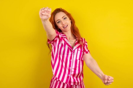 Foto de Mujer pelirroja con pijama rosa sobre fondo de estudio amarillo imaginar timón timón timón pasar examen de conducción buen humor velocidad rápida - Imagen libre de derechos