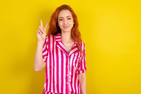 Photo for Red haired woman wearing pink pyjama over yellow studio background showing and pointing up with fingers number two while smiling confident and happy. - Royalty Free Image