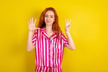 Foto de Mujer pelirroja vistiendo pijama rosa sobre fondo amarillo estudio mostrando y apuntando hacia arriba con los dedos número ocho mientras sonríe confiado y feliz. - Imagen libre de derechos