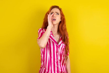 Foto de Mujer pelirroja vistiendo pijama rosa sobre fondo amarillo del estudio gritando y gritando fuerte a un lado con la mano en la boca. Concepto de comunicación. - Imagen libre de derechos
