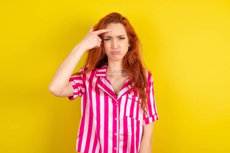 Photo for Red haired woman wearing pink pyjama over yellow studio background pointing unhappy at pimple on forehead, blackhead  infection. Skincare concept. - Royalty Free Image