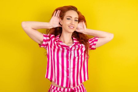 Foto de Mujer pelirroja vistiendo pijama rosa sobre fondo amarillo estudio tratando de escuchar ambas manos en el gesto de la oreja, curioso por chismes. Problema de audición, sordos - Imagen libre de derechos