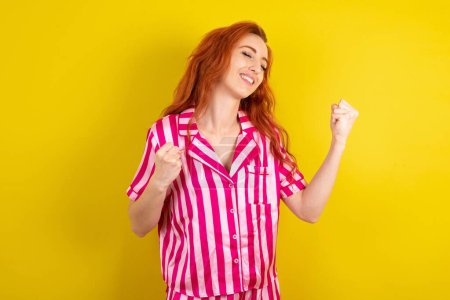 Téléchargez les photos : Femme aux cheveux roux portant un pyjama rose sur fond jaune studio célébrant surpris et étonnés du succès avec les bras levés et les yeux fermés. Concept gagnant. - en image libre de droit