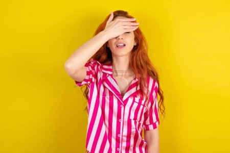 Photo for Red haired woman wearing pink pyjama over yellow studio background smiling and laughing with hand on face covering eyes for surprise. Blind concept. - Royalty Free Image