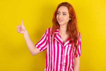Foto de Mujer pelirroja vistiendo pijama rosa sobre fondo amarillo estudio buscando orgulloso, sonriendo haciendo pulgares hacia arriba gesto a un lado. Buen trabajo.! - Imagen libre de derechos