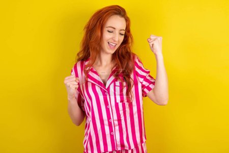 Téléchargez les photos : Femme aux cheveux rouges extatiques portant pyjama rose sur fond jaune studio crier fort oui poing levé gagner loterie - en image libre de droit