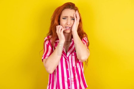 Photo for Doleful red haired woman wearing pink pyjama over yellow studio background crying woman, looks stressfully, frowns face, feels lonely and anxious - Royalty Free Image