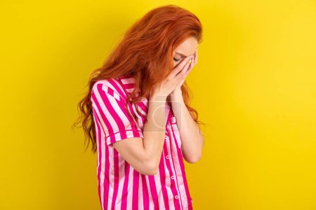 Foto de Triste mujer pelirroja vistiendo pijama rosa sobre fondo amarillo del estudio llorando cubriendo su cara con sus manos. - Imagen libre de derechos