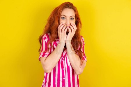 Téléchargez les photos : Vivace femme aux cheveux rouges portant pyjama rose sur fond jaune studio glousse et joyeusement, couvre la bouche, a rire naturel, entend une histoire positive ou anecdote drôle - en image libre de droit